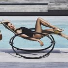 Mulher de maiô preto reclinada em uma cadeira de sol moderna, com sapatos plataforma dourados e uma bolsa marrom. Ela está à beira de uma piscina em um ambiente ensolarado e luxuoso.