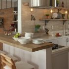 Cozinha moderna e aconchegante com ilha de madeira e balcão para refeições. Destaque para o revestimento de tijolinho, prateleiras abertas e luminárias pendentes, que criam um ambiente rústico e industrial.