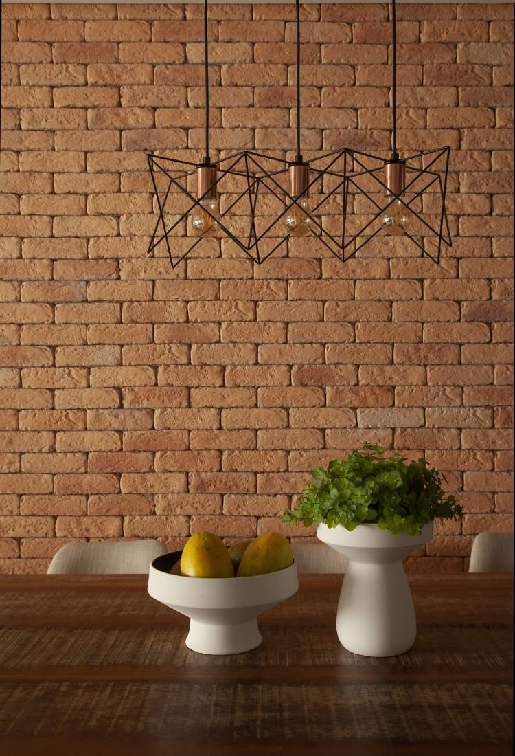 Sala de jantar com parede de tijolinhos aparentes e uma luminária pendente de metal com design geométrico. Sobre a mesa de madeira rústica, um vaso branco com plantas e uma fruteira com limões.