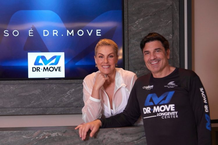 Dois atletas posam para uma foto dentro de uma máquina branca e aerodinâmica de alta tecnologia. A mulher loira sorri, enquanto um homem de cabelo escuro, com uma camiseta preta da DR MOVE, coloca o braço em seu ombro. A máquina tem a marca "MOVE SPACE" e a bandeira do Brasil.