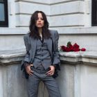 Jenna Ortega veste um terno de três peças cinza. O casaco tem ombros pontudos e as mangas de renda preta saem das mangas do casaco. Ela usa uma colete e calças combinando. Um buquê de rosas vermelhas está ao lado dela.