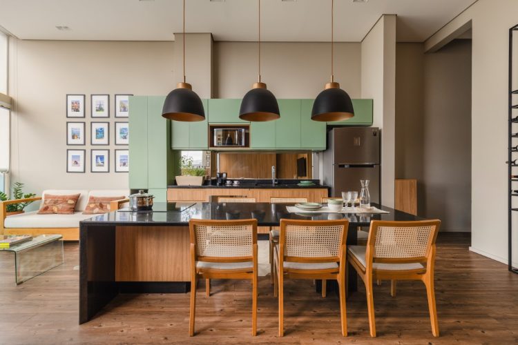 Cozinha moderna e sala de estar em plano aberto. A cozinha tem armários superiores em verde-claro, armários inferiores de madeira, e uma ilha preta com cadeiras de vime e madeira. Acima da ilha, três luminárias pendentes pretas. A sala de estar, ao fundo, tem um sofá branco e quadros decorativos na parede.