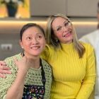 Duas mulheres sorriem para a câmera, com uma delas, Gloria, a abraçando. Ao fundo, um homem desfocado com roupa de chef de cozinha sorri. A foto foi tirada em uma cozinha.
