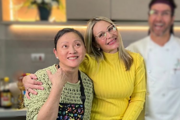 Duas mulheres sorriem para a câmera, com uma delas, Gloria, a abraçando. Ao fundo, um homem desfocado com roupa de chef de cozinha sorri. A foto foi tirada em uma cozinha.