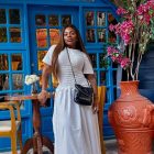 Jojo Todynho, em um vestido branco e bolsa preta Chanel, posa em uma área externa. Ao fundo, uma porta azul, um vaso de barro com um arranjo floral rosa e um vaso com limões amarelos.