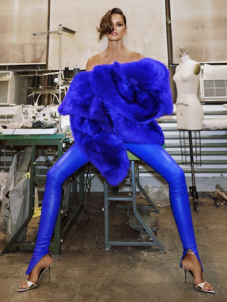Izabel Goulart usando salto fino, calça azul e máxi casaco azul, sentada sobre cadeira em ateliê