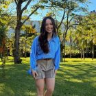Uma mulher sorridente com cabelos longos e escuros, vestindo uma camisa azul listrada e shorts bege, posa em um jardim ensolarado. O fundo é composto por árvores, gramado verde e uma piscina, com um prédio visível ao longe.