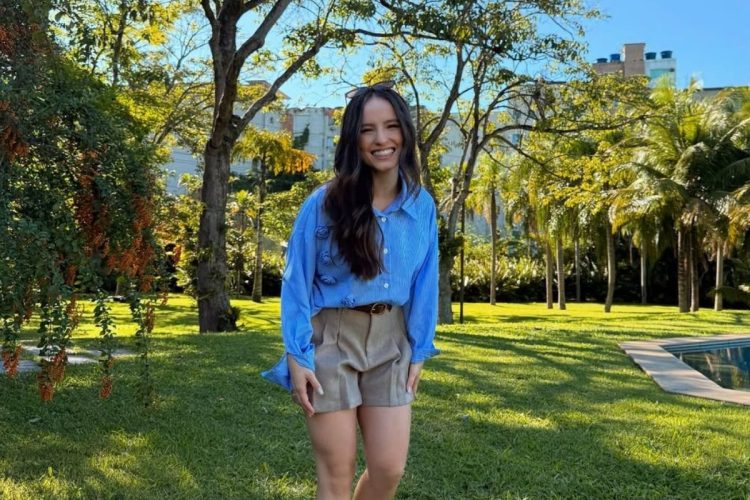 Uma mulher sorridente com cabelos longos e escuros, vestindo uma camisa azul listrada e shorts bege, posa em um jardim ensolarado. O fundo é composto por árvores, gramado verde e uma piscina, com um prédio visível ao longe.