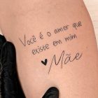 A imagem mostra uma tatuagem no braço de uma pessoa com os dizeres "Você é o amor que existe em mim ❤️ Mãe". A tatuagem é escrita em fonte cursiva, com um pequeno coração antes da palavra "Mãe". A pele ao redor da tatuagem parece clara e lisa, e uma mão enluvada de preto segura o braço.