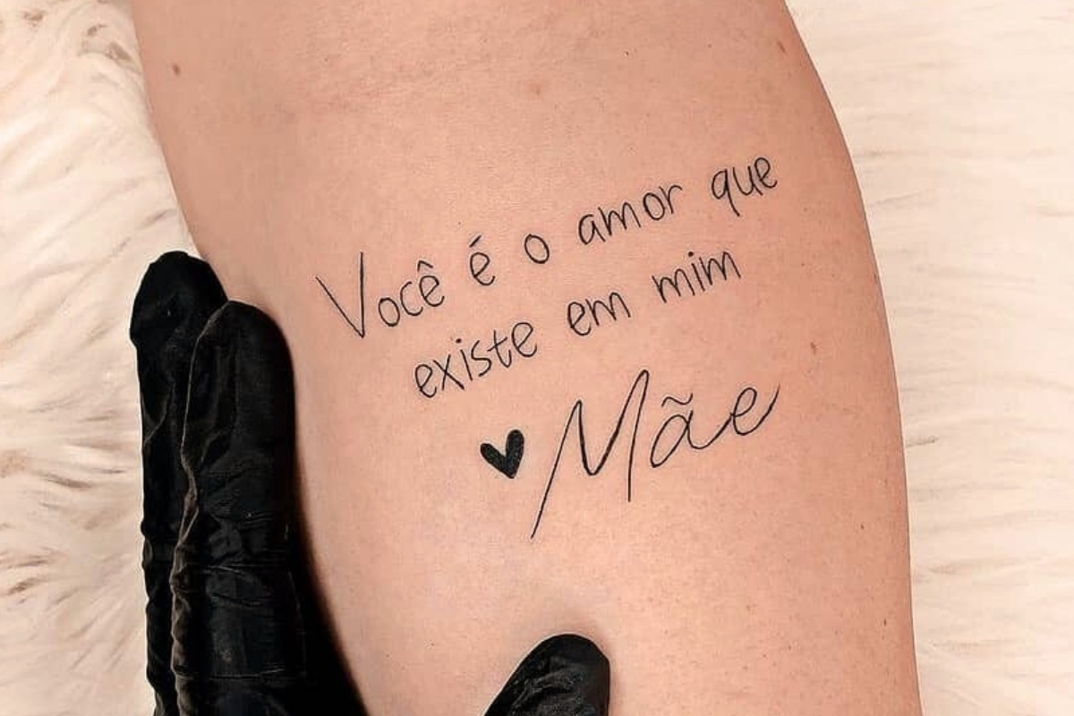 A imagem mostra uma tatuagem no braço de uma pessoa com os dizeres "Você é o amor que existe em mim ❤️ Mãe". A tatuagem é escrita em fonte cursiva, com um pequeno coração antes da palavra "Mãe". A pele ao redor da tatuagem parece clara e lisa, e uma mão enluvada de preto segura o braço.