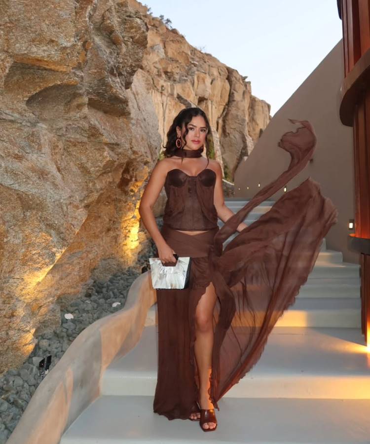 Maisa Silva em um elegante vestido de seda marrom posa em uma escadaria externa. A parte de cima é um espartilho, enquanto a saia possui uma fenda e tecido esvoaçante. Ao fundo, uma parede de rocha com iluminação e uma estrutura arquitetônica.
