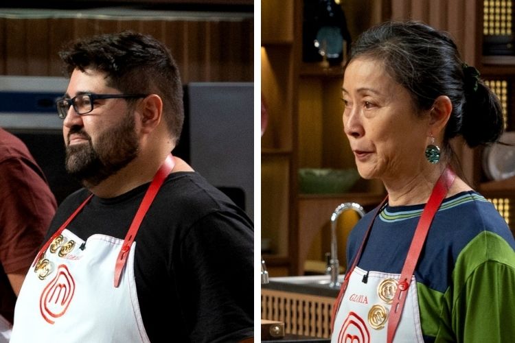 Homem de óculos e barba, e mulher com coque, ambos vestindo aventais de chef brancos com detalhes em vermelho. Eles estão em uma cozinha, olhando para a direita da imagem. O homem está de perfil, e a mulher está de lado.