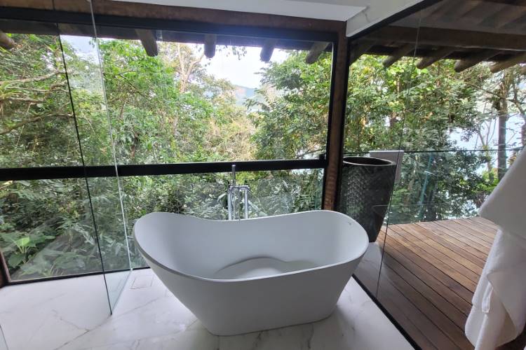 Banheira moderna em um banheiro com vista panorâmica para a mata exuberante. O espaço combina elementos rústicos, como a madeira, com toques de luxo e janelas que integram o ambiente à natureza.