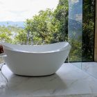 Banheira moderna e elegante em um banheiro luxuoso com janelas grandes, vista para a natureza exuberante e montanhas ao longe. O piso é de mármore claro e há uma mesa lateral de vidro.