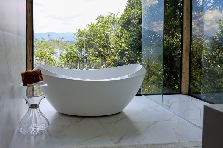 Banheira moderna e elegante em um banheiro luxuoso com janelas grandes, vista para a natureza exuberante e montanhas ao longe. O piso é de mármore claro e há uma mesa lateral de vidro.