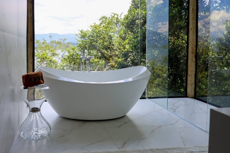 Banheira moderna e elegante em um banheiro luxuoso com janelas grandes, vista para a natureza exuberante e montanhas ao longe. O piso é de mármore claro e há uma mesa lateral de vidro.