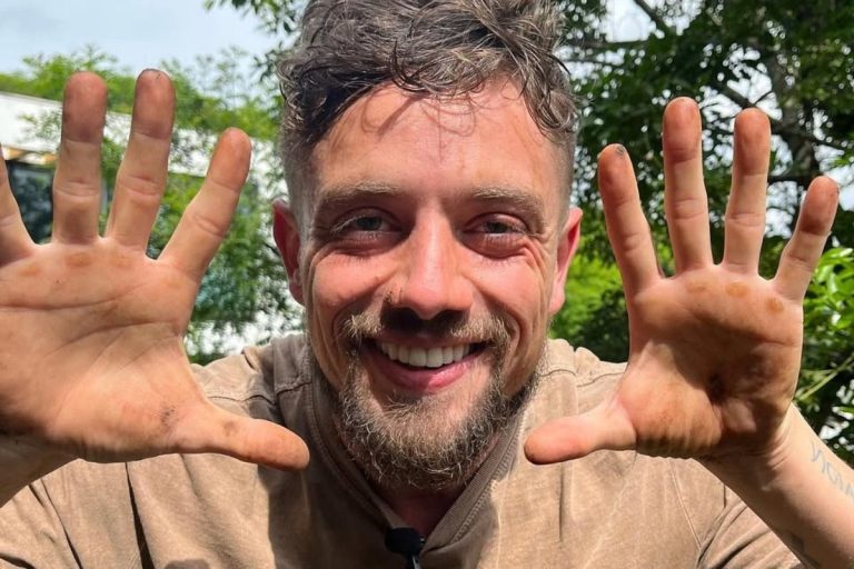 Um homem com barba e cabelos castanhos, vestindo uma camiseta marrom, sorri e mostra as mãos sujas de terra para a câmera. Ele está em um ambiente externo com vegetação ao fundo.