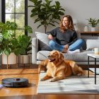 Uma mulher sorridente sentada no sofá acaricia um golden retriever deitado. Um robô aspirador de pó preto limpa o chão de madeira, em uma sala de estar clara e moderna com janelas grandes e muitas plantas.