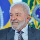 Presidente Luiz Inácio Lula da Silva sorri levemente, olhando para a direita. Ele tem cabelos e barba grisalhos, vestindo um terno cinza e uma gravata azul listrada. A bandeira do Brasil está desfocada ao fundo.