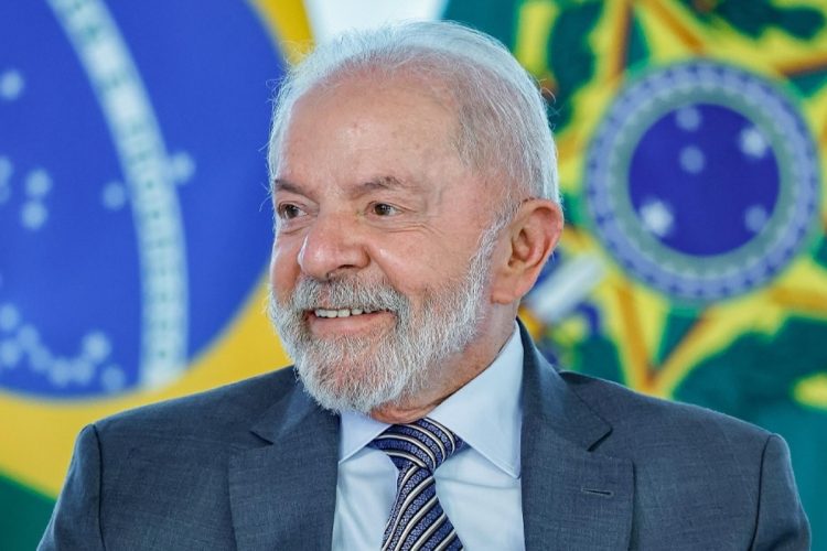 Presidente Luiz Inácio Lula da Silva sorri levemente, olhando para a direita. Ele tem cabelos e barba grisalhos, vestindo um terno cinza e uma gravata azul listrada. A bandeira do Brasil está desfocada ao fundo.