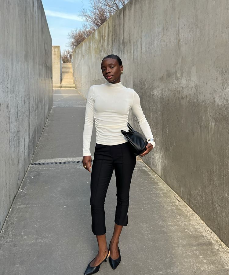 Mulher com blusa branca de gola alta e calça preta. Ela está em um corredor de concreto, segurando uma bolsa preta. A imagem mostra seu corpo inteiro e um ambiente minimalista.