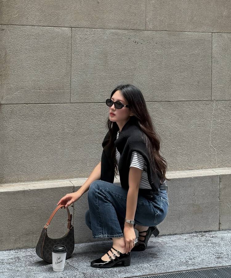 Mulher agachada em frente a uma parede de pedra, usando óculos de sol, camiseta listrada, calça jeans, suéter preto amarrado nos ombros e **sapato preto** de fivela. Há um copo de café e uma bolsa próxima a ela.