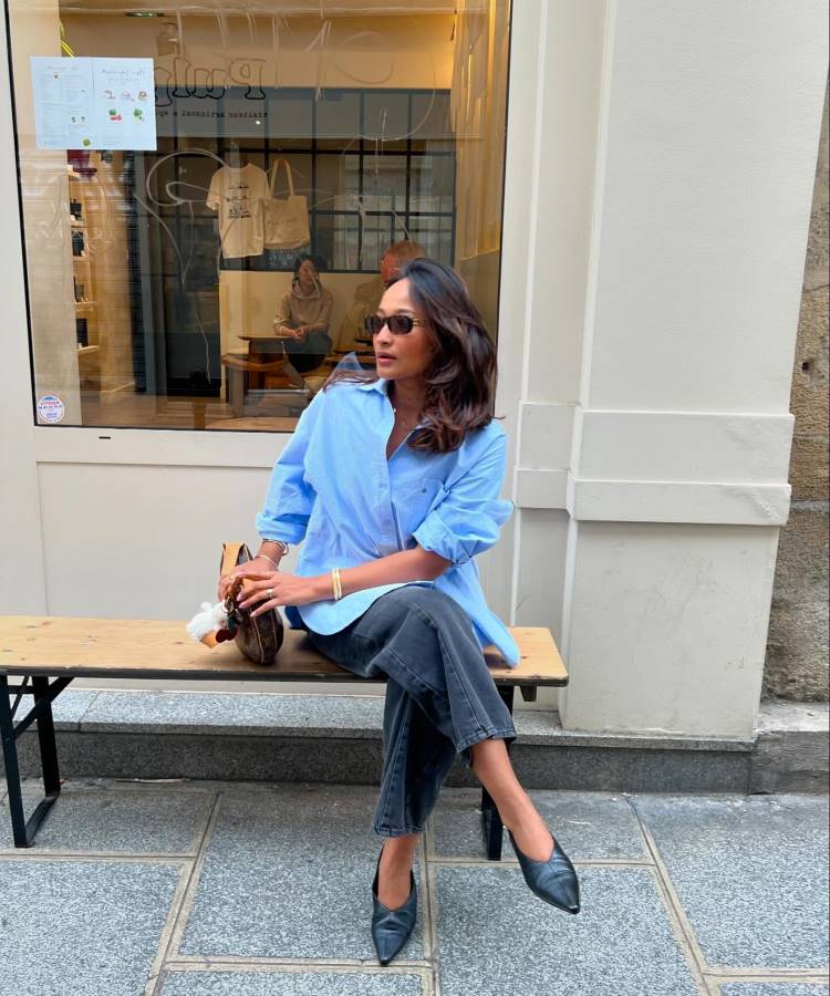 Mulher sentada em um banco de madeira, vestindo uma camisa azul, calça jeans preta e sapatos de salto pretos. Ela usa óculos escuros, segura uma bolsa e posa em frente a uma vitrine de loja em um ambiente urbano.
