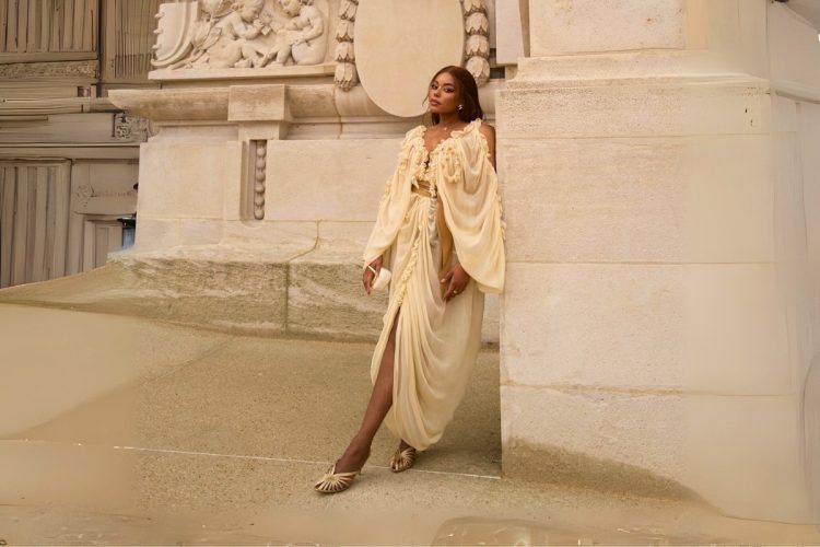 Uma mulher posa elegantemente em frente a um edifício de pedra. Ela usa um vestido creme esvoaçante com mangas compridas e um cinto dourado, sapatos dourados e brincos grandes, olhando para longe da câmera.
