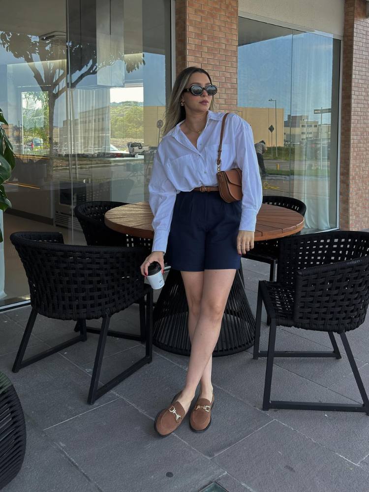 Mulher loira de pele clara usando camisa branca, shorts de alfaiataria preto e mocassim marrom 