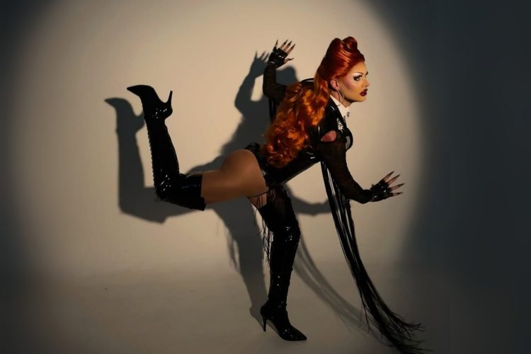 Artista em estúdio iluminado dramaticamente com feixe de luz. Usa body preto recortado, franjas longas e botas de verniz. Cabelos ruivos volumosos caem sobre os ombros. Corpo inclinado em pose performática, expressando energia e intensidade de apresentação drag estilizada.