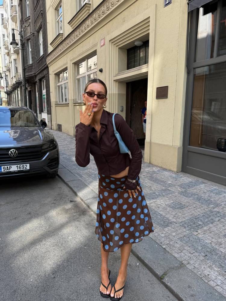 Mulher de pele clara de camisa marrom, saia midi marrom transparente com poá azul e bolsa azul