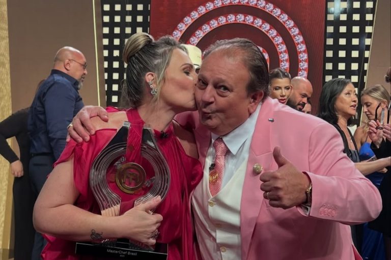 A chef Daniela Dantas, com o troféu do MasterChef Brasil 2025, abraça e beija um homem de terno rosa. Ele está de perfil, dando um sinal de positivo com o polegar.