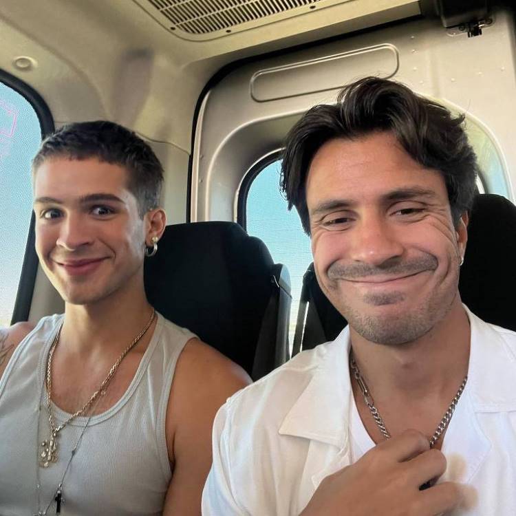 Dois homens tiram uma selfie dentro de um veículo. O homem à esquerda sorri para a câmera, enquanto o da direita faz uma careta e aperta a gola de sua camisa branca. famosos que são parentes