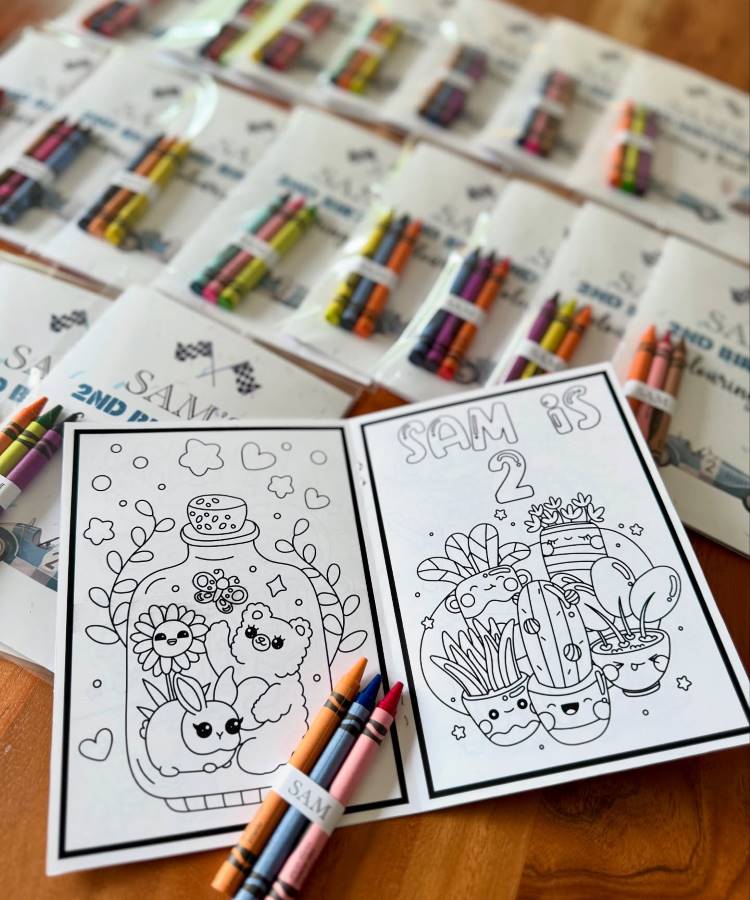 Livros de colorir personalizados com o nome "SAM" e a idade "2". Vários kits de giz de cera, cada um com quatro cores, estão dispostos ao redor, embalados e prontos para serem dados como lembrancinhas.
