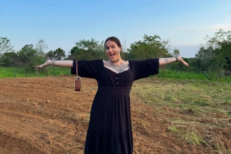 Pessoa com vestido preto longo posa sorridente em campo aberto de terra e vegetação. De braços abertos, transmite energia alegre e descontraída. Tatuagens visíveis no peito e braços. O céu azul e árvores ao fundo completam a cena natural.