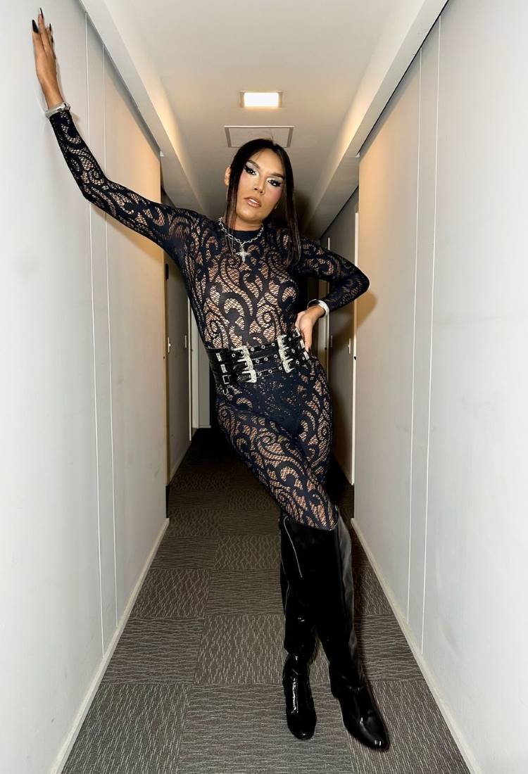 Uma pessoa alta e magra com cabelo liso e preto, usando um macacão de renda preta e botas de cano alto da mesma cor. Ela está apoiada na parede com uma mão, em um corredor estreito.