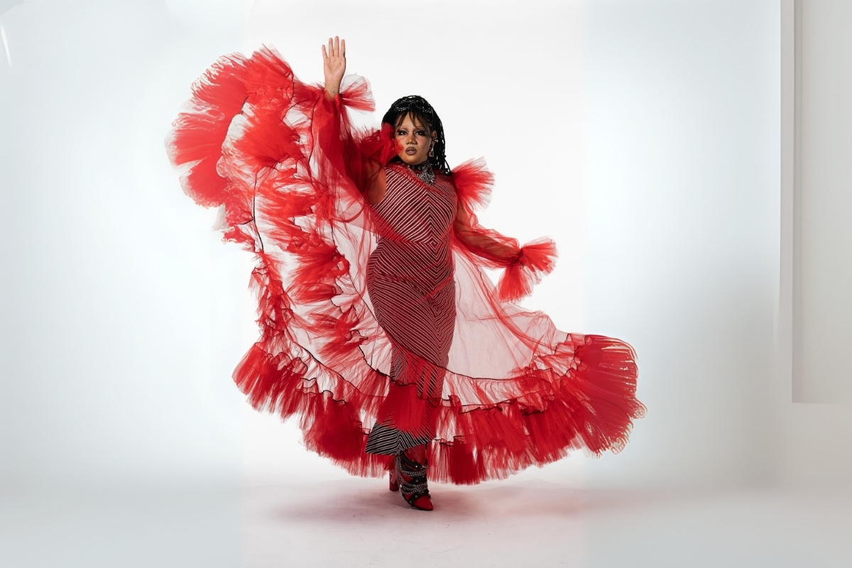 Uma pessoa transfeminina deslumbrante em um vestido longo listrado vermelho e branco, com mangas e cauda exuberantes de tule vermelho esvoaçante, posando com um braço levantado. Ela tem maquiagem dramática e usa sapatos de salto alto vermelhos e brilhantes.