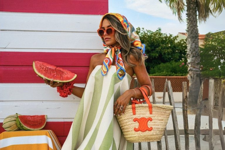 Mulher em frente a uma barraca de praia com estampa de listras vermelhas e brancas, usando um vestido listrado verticalmente nas cores creme e verde, um lenço de seda na cabeça, óculos escuros, e segurando uma fatia de melancia e uma bolsa de palha da Céline.