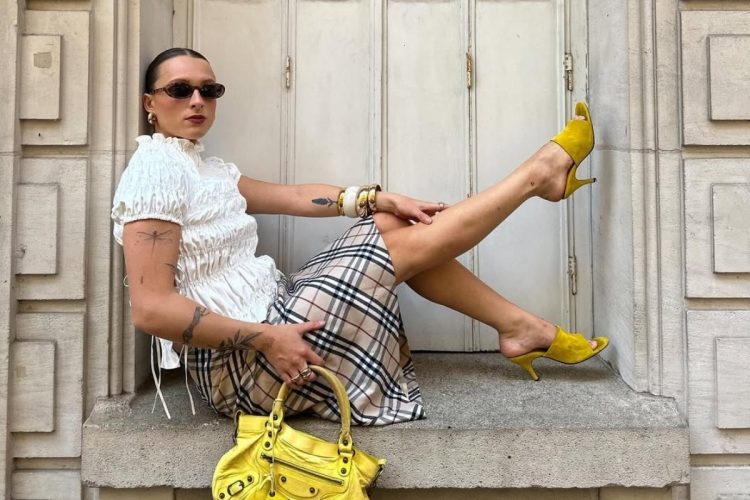 Foto de influencer usando óculos de sol, blusa branca, saia xadrez e tamanco amarelo, uma das tendências de sandálias para o verão 2026