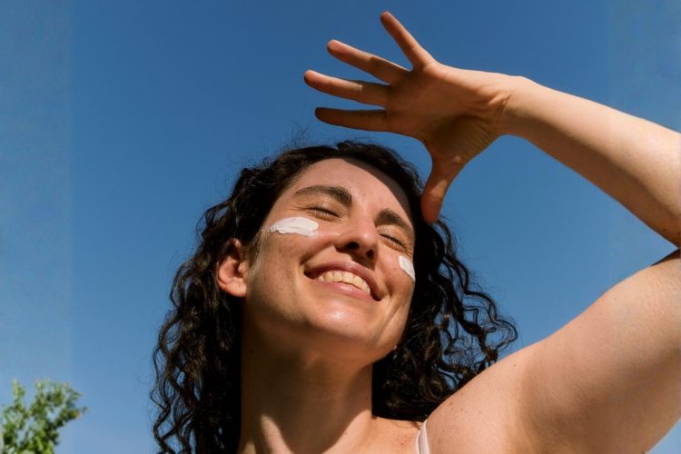 Foto de mulher usando protetor solar facial e colocando a mão no rosto para se proteger do sol