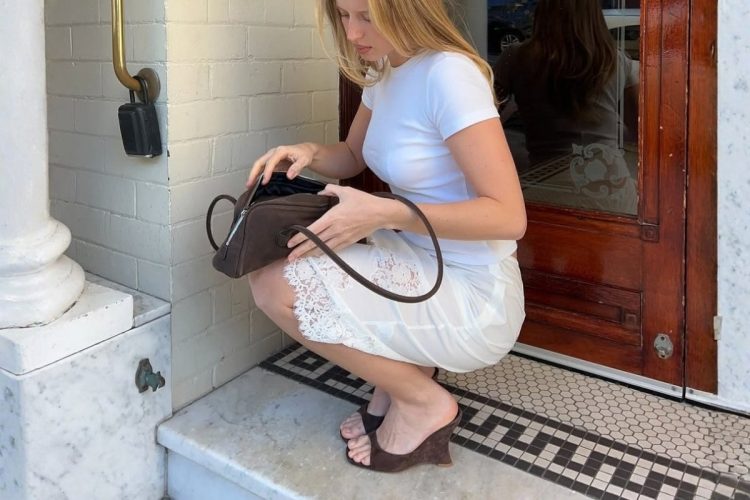 Foto de mulher loira agachada usando camiseta e saia mídi brancas com tamanco marrom de salto anabela