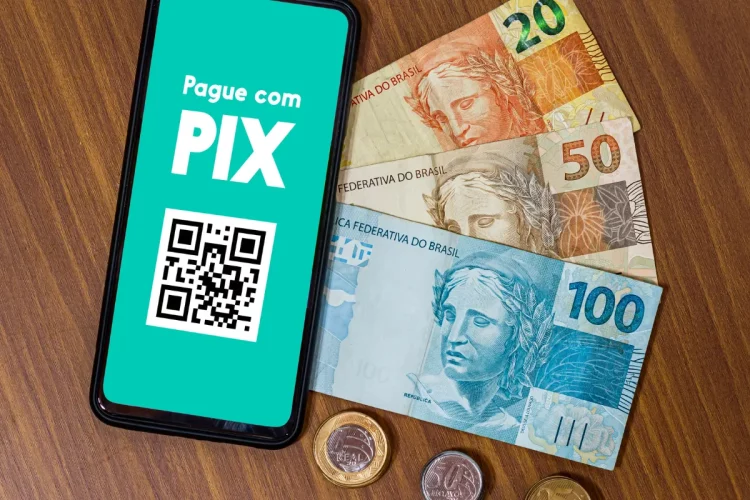 Celular com tela de pagamento Pix ao lado de notas e moedas de real sobre mesa de madeira