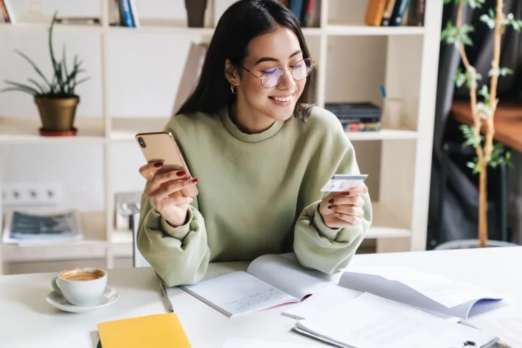 Mulher usando celular e cartão de crédito em casa enquanto compara opções de cartão sem anuidade em 2026.