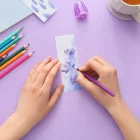 Pessoa desenhando marcador de páginas floral com lápis coloridos sobre mesa lilás em tutorial criativo.