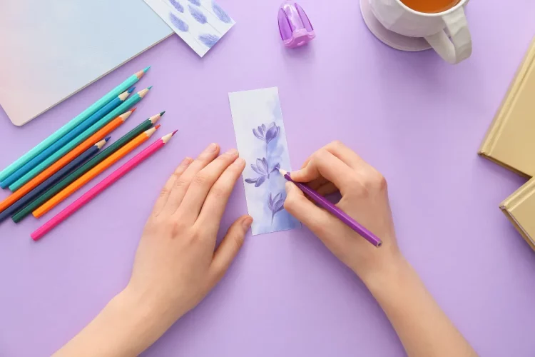 Pessoa desenhando marcador de páginas floral com lápis coloridos sobre mesa lilás em tutorial criativo.