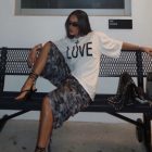 Foto de mulher morena sentada em banco usando camiseta branca, bermuda com estampa camuflada e chinelo de salto preto