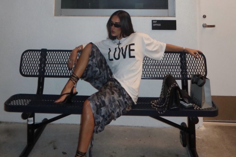 Foto de mulher morena sentada em banco usando camiseta branca, bermuda com estampa camuflada e chinelo de salto preto