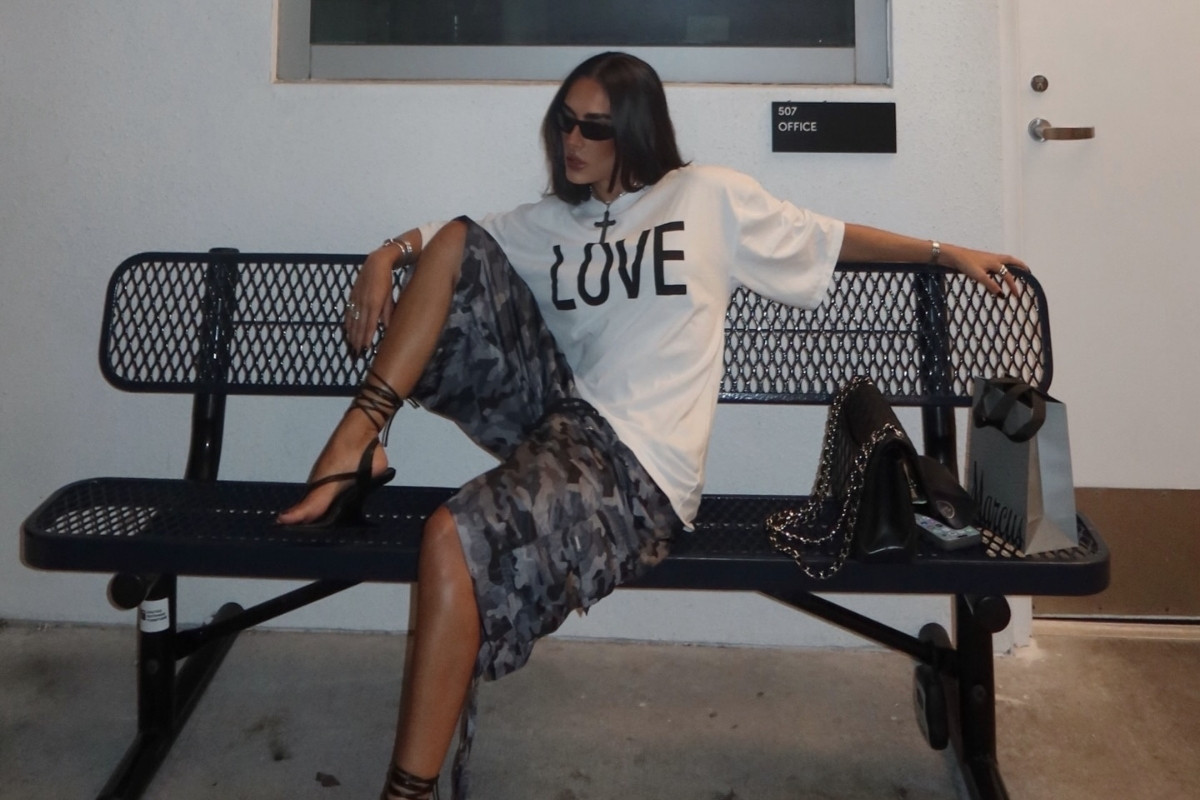Foto de mulher morena sentada em banco usando camiseta branca, bermuda com estampa camuflada e chinelo de salto preto