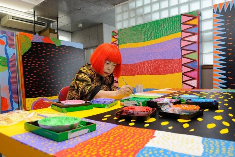 Foto de Yayoi Kusama de cabelo vermelho sentada em uma mesa pintando